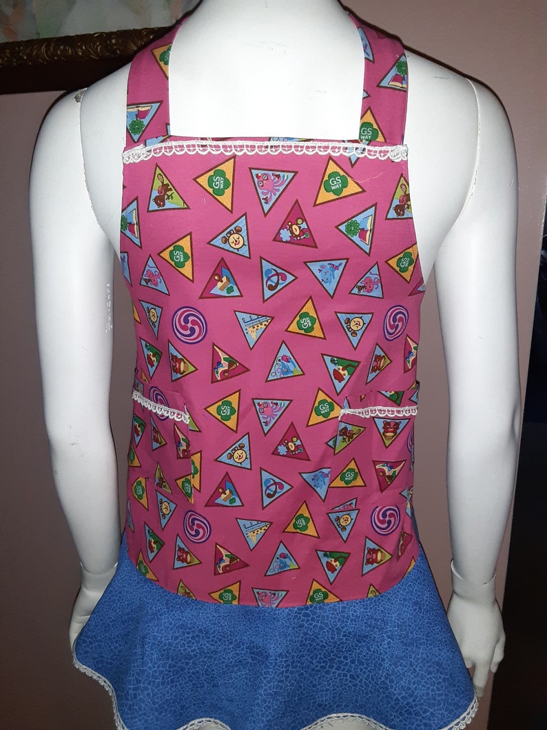 May include: Pink and blue Girl Scout apron with a white lace trim. The apron has a pattern of colorful triangles with Girl Scout badges and symbols. The apron has two pockets.