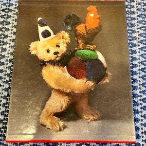 May include: A brown teddy bear wearing a white and blue hat holds a multi-colored ball. A red rooster with a black and white tail sits on top of the ball.
