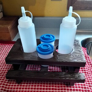 Pode incluir: Um suporte para condimentos em estilo piquenique marrom escuro com duas garrafas transparentes e dois recipientes pequenos com tampas azuis. O suporte está sobre uma toalha de mesa xadrez vermelha e branca.