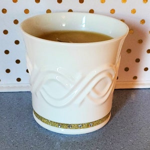 May include: A white ceramic candle holder with a gold glitter band and rhinestones. The candle holder has a decorative embossed design and contains a light brown candle. The candle holder is on a gray surface.