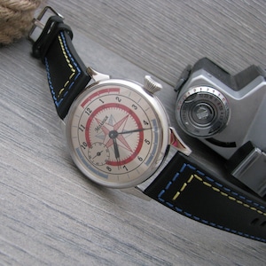 Può includere: Un orologio da polso argentato con quadrante bianco e dettagli rossi. L'orologio ha un cinturino in pelle nera con cuciture blu e gialle. Il quadrante dell'orologio presenta una rosa dei venti e la parola "Молния" in scrittura cirillica.