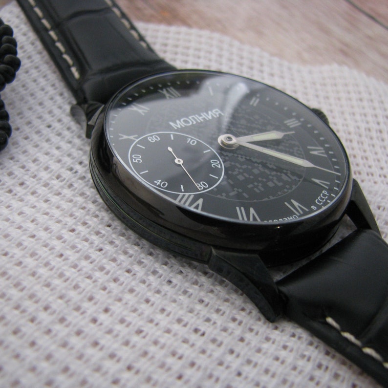 May include: A black wristwatch with a black leather band and a white face. The watch face has Roman numeral hour markers and a small seconds dial. The watch is made in the USSR.