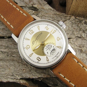 Puede incluir: Un reloj de pulsera plateado con una esfera blanca y detalles dorados. El reloj tiene una correa de cuero marr&oacute;n con costuras blancas. La esfera del reloj tiene las palabras "Спутник" y "СССР" impresas.