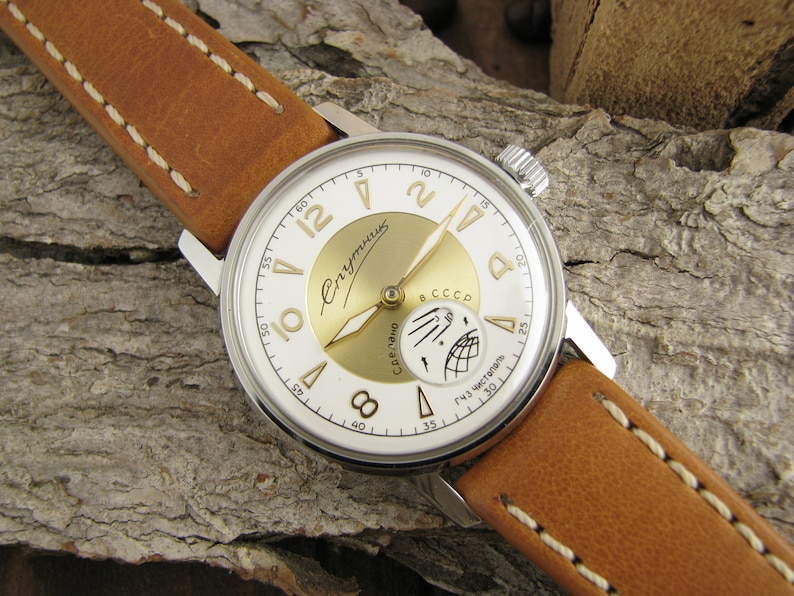 Puede incluir: Un reloj de pulsera plateado con una esfera blanca y detalles dorados. El reloj tiene una correa de cuero marr&oacute;n con costuras blancas. La esfera del reloj tiene las palabras "Спутник" y "СССР" impresas.