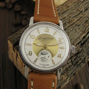 Puede incluir: Un reloj de pulsera plateado con una esfera blanca y detalles dorados. La esfera del reloj tiene el texto "Спутник" y "Сделано в СССР". El reloj tiene una correa de cuero marr&oacute;n.