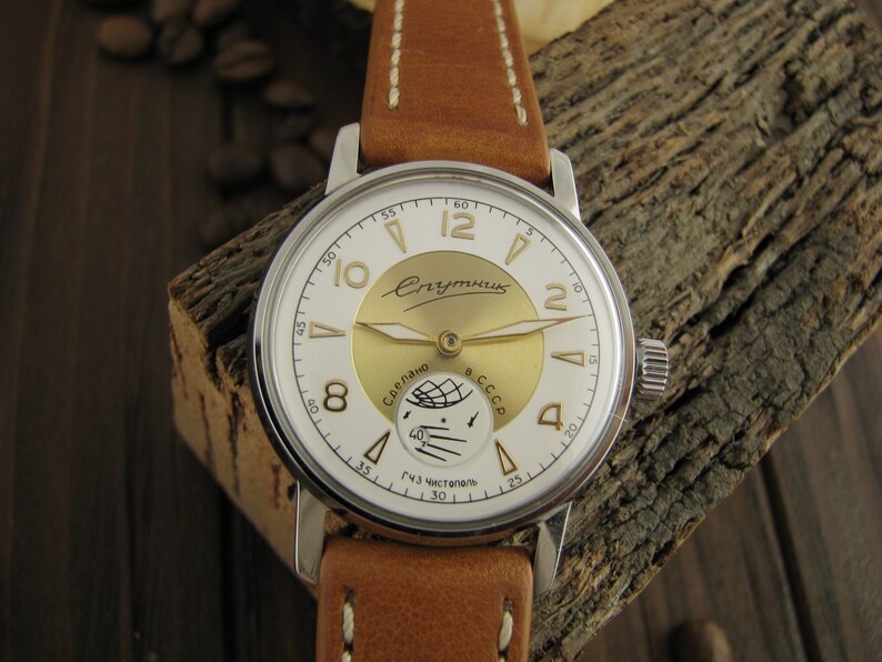 Puede incluir: Un reloj de pulsera plateado con una esfera blanca y detalles dorados. La esfera del reloj tiene el texto "Спутник" y "Сделано в СССР". El reloj tiene una correa de cuero marr&oacute;n.