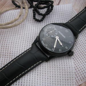 May include: A black wristwatch with a black leather band and white stitching. The watch face is black with white Roman numeral hour markers and a white second hand. The watch face also has the word "МОЛНИЯ" printed in white.