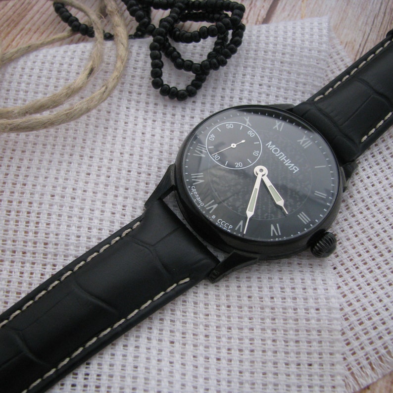 May include: A black wristwatch with a black leather band and white stitching. The watch face is black with white Roman numeral hour markers and a white second hand. The watch face also has the word "МОЛНИЯ" printed in white.