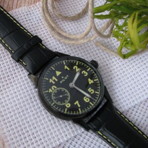 May include: A black pilot watch with a yellow face and black hands. The watch has a black leather strap with yellow stitching. The watch face has the word "PILOT" printed on it.