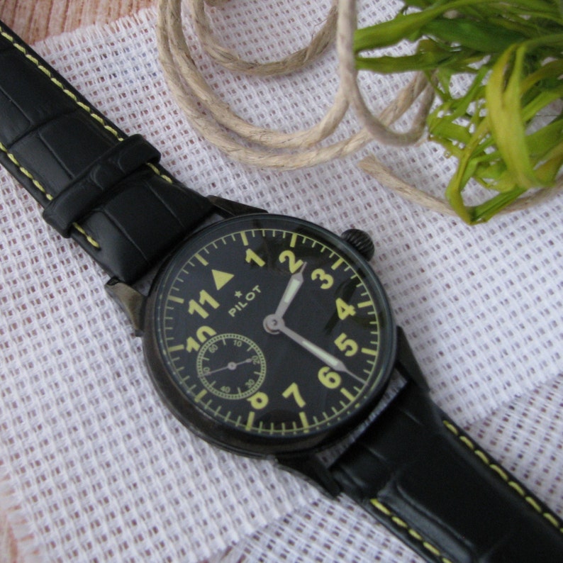 May include: A black pilot watch with a yellow face and black hands. The watch has a black leather strap with yellow stitching. The watch face has the word "PILOT" printed on it.