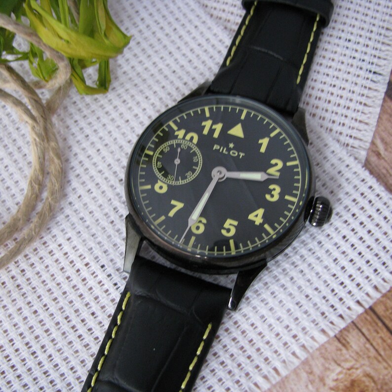 May include: Black leather wristwatch with a black face and yellow numerals. The watch has a small subdial and the word "PILOT" printed on the face.