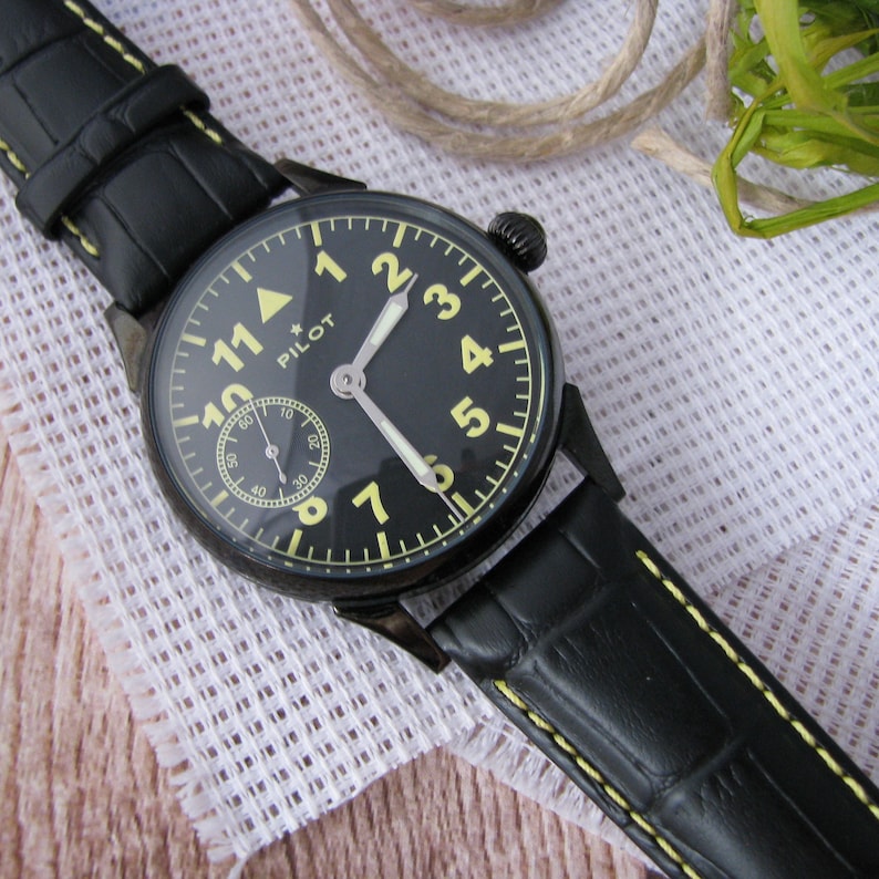 May include: A black pilot watch with a yellow face and black leather strap. The watch face has large white numbers and a small star above the word "PILOT".