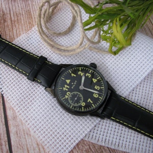 May include: A black leather watch with a yellow face and black hands. The watch face has white numbers and the word "PILOT" printed on it.
