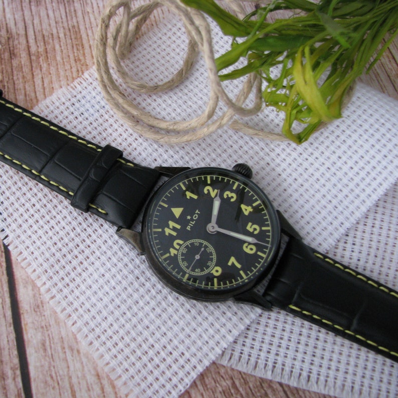 May include: A black leather watch with a yellow face and black hands. The watch face has white numbers and the word "PILOT" printed on it.