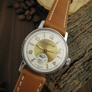 Puede incluir: Un reloj de pulsera plateado con una esfera blanca y detalles dorados. La esfera del reloj presenta una representaci&oacute;n de un sat&eacute;lite orbitando la Tierra, con el texto "Спутник" y "Сделано в СССР" en escritura cir&iacute;lica. El reloj tiene una correa de cuero marr&oacute;n.