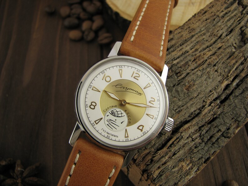Puede incluir: Un reloj de pulsera plateado con una esfera blanca y detalles dorados. La esfera del reloj presenta una representaci&oacute;n de un sat&eacute;lite orbitando la Tierra, con el texto "Спутник" y "Сделано в СССР" en escritura cir&iacute;lica. El reloj tiene una correa de cuero marr&oacute;n.