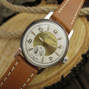 Puede incluir: Un reloj de pulsera plateado con una esfera blanca y detalles dorados. El reloj tiene una correa de cuero marr&oacute;n con costuras blancas. La esfera del reloj tiene las palabras "СССР" y "Сделано" en escritura cir&iacute;lica. El reloj est&aacute; sobre una superficie de madera.