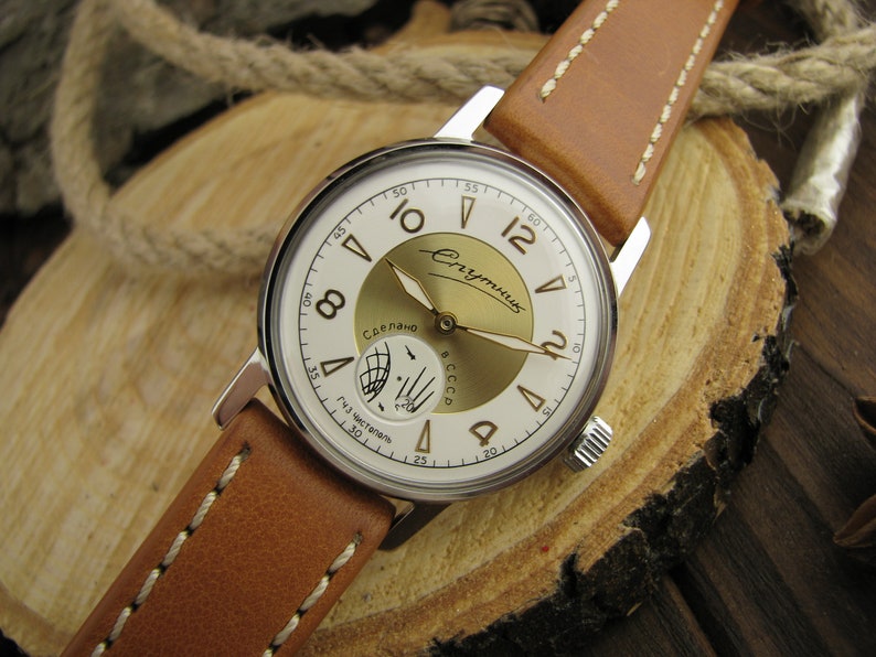 Puede incluir: Un reloj de pulsera plateado con una esfera blanca y detalles dorados. El reloj tiene una correa de cuero marr&oacute;n con costuras blancas. La esfera del reloj tiene las palabras "СССР" y "Сделано" en escritura cir&iacute;lica. El reloj est&aacute; sobre una superficie de madera.