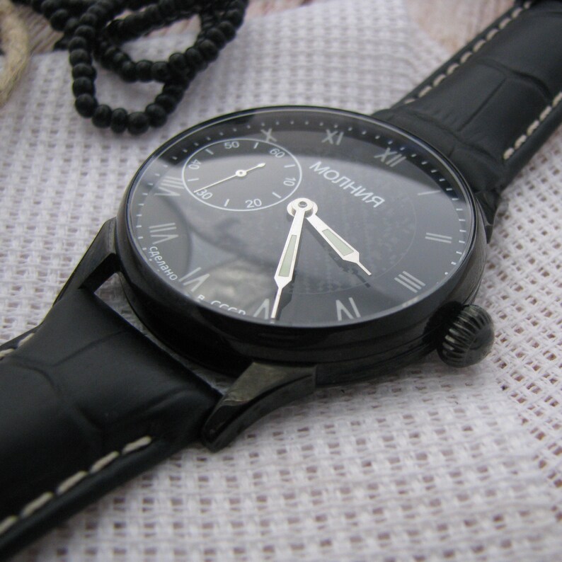 May include: A black wristwatch with a black leather band and a white face. The watch face has Roman numeral hour markers and a small seconds dial. The watch is labeled "Молния" which is Russian for "Lightning".