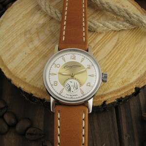 Puede incluir: Un reloj de pulsera plateado con una esfera blanca y detalles dorados. El reloj tiene una correa de cuero marr&oacute;n con costuras blancas. La esfera del reloj tiene las palabras "СССР" y "Чайка" impresas.