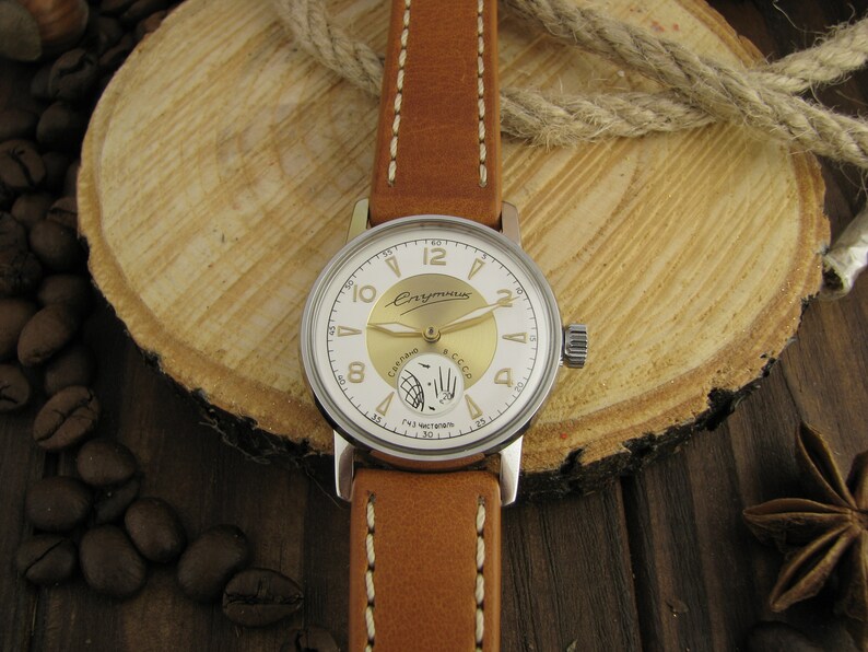 Puede incluir: Un reloj de pulsera plateado con una esfera blanca y detalles dorados. El reloj tiene una correa de cuero marr&oacute;n con costuras blancas. La esfera del reloj tiene las palabras "СССР" y "Чайка" impresas.
