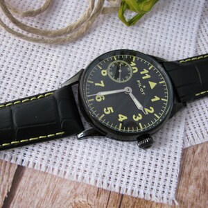 May include: A black leather wristwatch with yellow stitching. The watch face is black with yellow numerals and a small white subdial. The watch face also has the word "PILOT" printed on it.