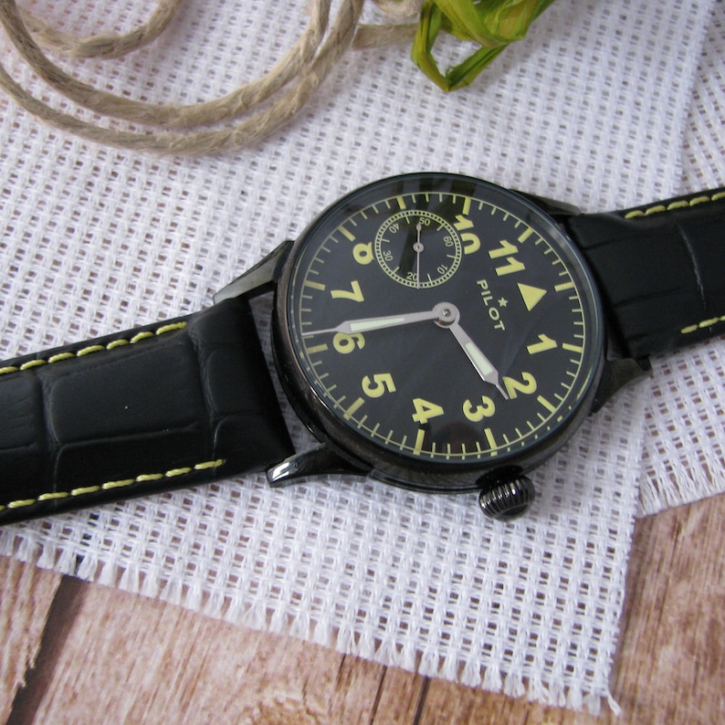 May include: A black leather wristwatch with yellow stitching. The watch face is black with yellow numerals and a small white subdial. The watch face also has the word "PILOT" printed on it.