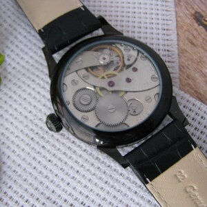 May include: A black wristwatch with a leather band and a clear caseback revealing the mechanical movement. The watch has a silver face and black hands.
