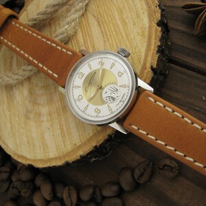 Puede incluir: Un reloj de pulsera plateado con una esfera blanca y detalles dorados. El reloj tiene una correa de cuero marr&oacute;n con costuras blancas. La esfera del reloj tiene la palabra "Слава" en escritura cir&iacute;lica y las palabras "Made in USSR" en ingl&eacute;s.