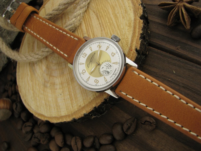 Puede incluir: Un reloj de pulsera plateado con una esfera blanca y detalles dorados. El reloj tiene una correa de cuero marr&oacute;n con costuras blancas. La esfera del reloj tiene la palabra "Слава" en escritura cir&iacute;lica y las palabras "Made in USSR" en ingl&eacute;s.