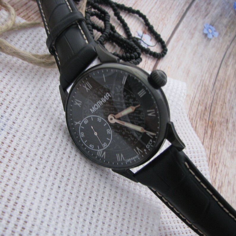 May include: A black wristwatch with a black leather band and a white face. The face has Roman numeral hour markers and the words "Молния" and "СССР" printed on it.