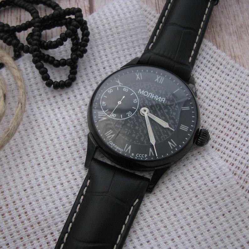 May include: A black wristwatch with a black leather band and a white face. The watch face has Roman numeral hour markers and a small seconds dial. The watch face also has the word "МОЛНИЯ" printed on it, which is Russian for "Lightning".