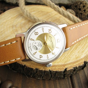 Puede incluir: Un reloj de pulsera plateado con una esfera blanca y detalles dorados. El reloj tiene una correa de cuero marr&oacute;n y presenta el texto "Сделано в СССР" y "Чайка" en la esfera.