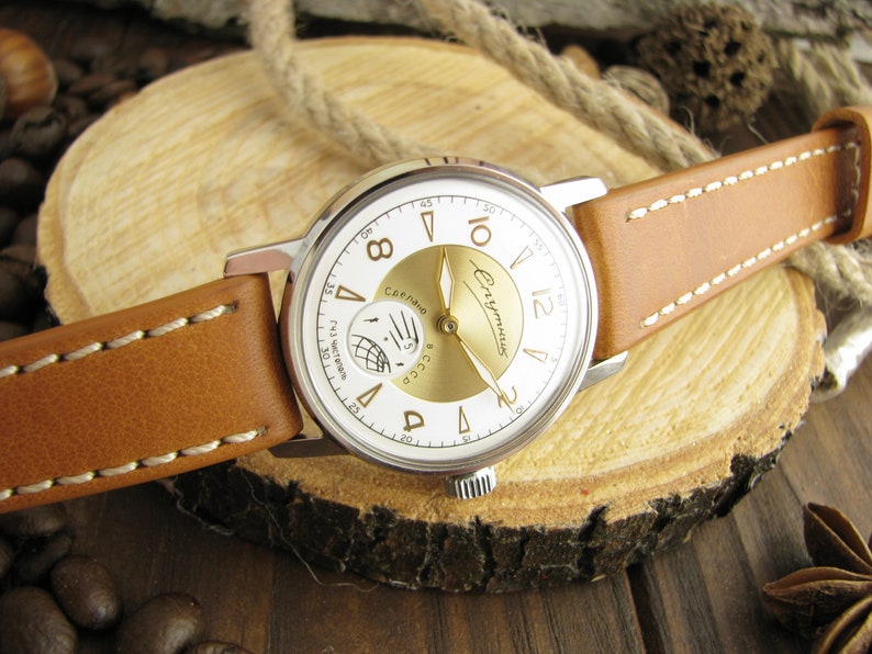 Puede incluir: Un reloj de pulsera plateado con una esfera blanca y detalles dorados. El reloj tiene una correa de cuero marr&oacute;n y presenta el texto "Сделано в СССР" y "Чайка" en la esfera.
