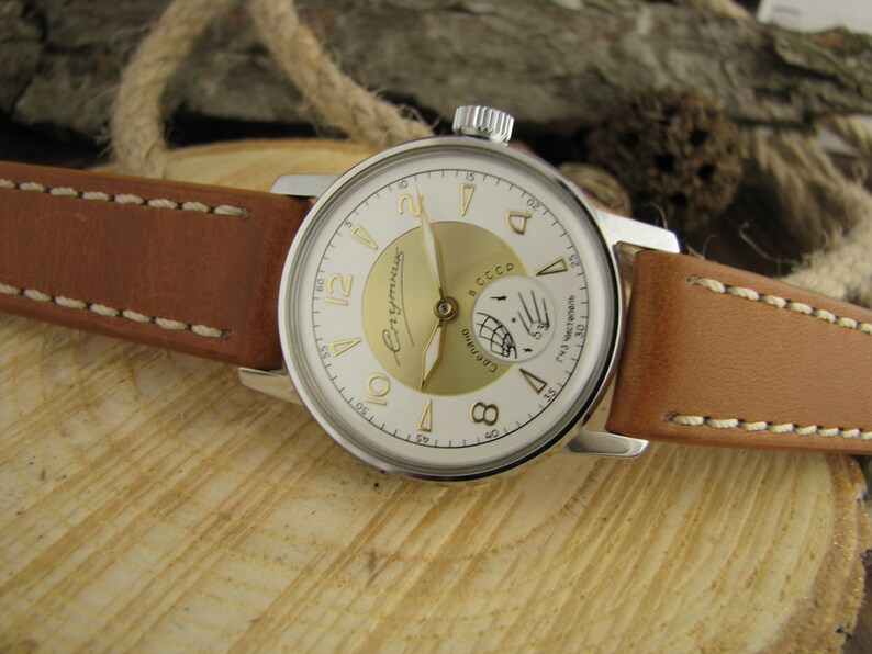 Puede incluir: Un reloj de pulsera plateado con una esfera blanca y detalles dorados. El reloj tiene una correa de cuero marr&oacute;n con costuras blancas. La esfera del reloj tiene las palabras "СССР" y "ЧaconoB" impresas.