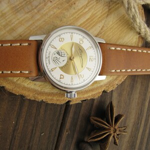 Puede incluir: Un reloj de pulsera plateado con una esfera blanca y detalles dorados. El reloj tiene una correa de cuero marr&oacute;n con costuras blancas. La esfera del reloj tiene el texto "СССР" y "ЧacOBbI" en escritura cir&iacute;lica.