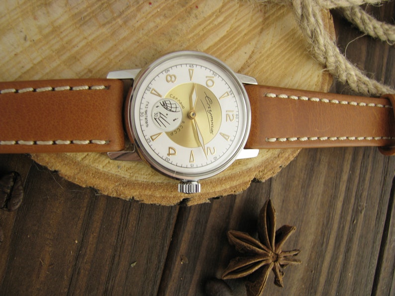 Puede incluir: Un reloj de pulsera plateado con una esfera blanca y detalles dorados. El reloj tiene una correa de cuero marr&oacute;n con costuras blancas. La esfera del reloj tiene el texto "СССР" y "ЧacOBbI" en escritura cir&iacute;lica.