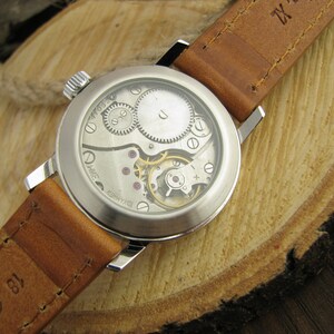 Puede incluir: Un reloj de pulsera plateado con una correa de cuero marr&oacute;n. El reloj tiene una parte posterior transparente que revela el funcionamiento interno del reloj.