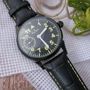 May include: A black pilot watch with a black leather strap and yellow stitching. The watch face has white hands and yellow numbers. The watch face also has a small subdial at the 3 o'clock position. The watch face says "PILOT" at the 12 o'clock position.