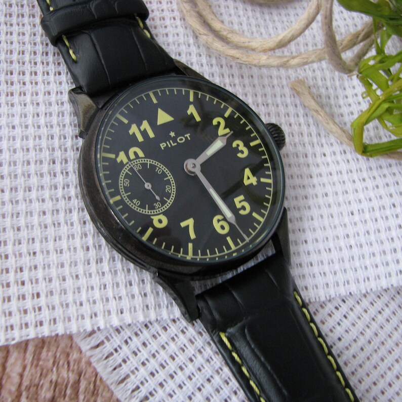 May include: A black pilot watch with a black leather strap and yellow stitching. The watch face has white numbers and hands, and a small subdial with a white hand. The watch face also has the word "PILOT" printed on it.