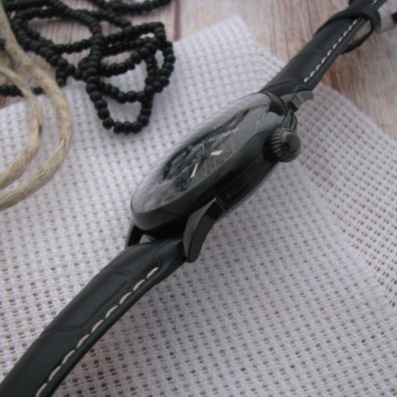 May include: A black wristwatch with a black leather band and white stitching. The watch face is black with white markings.