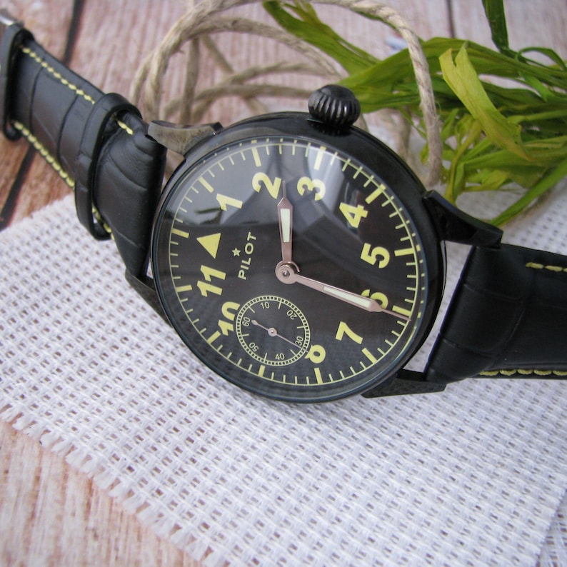 May include: A black pilot watch with a yellow face and black leather band. The watch face has large white numbers and a small white star at the top. The watch face also has a small second hand.
