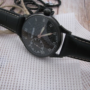 May include: A black wristwatch with a black leather band. The watch face is black with white Roman numeral hour markers and a white second hand. The watch face also has the word "Молния" in white letters and the text "Сделано в СССР" in white letters.
