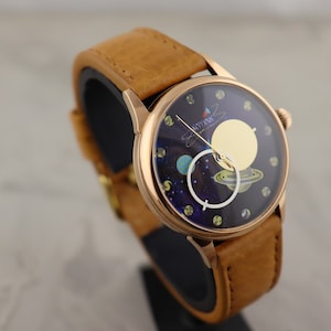 May include: A gold-tone wristwatch with a brown leather band. The watch face features a black background with a depiction of the solar system, including the sun, planets, and stars.