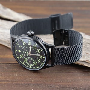 May include: A black pilot watch with a black mesh strap. The watch face has a black background with white numbers and a green second hand. The watch face also has the word "PILOT" printed on it.