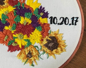 May include: Embroidered hoop art featuring a bouquet of sunflowers, daisies, and other flowers in shades of yellow, orange, red, purple, and green. The date "10.20.17" is embroidered in black thread on the white fabric.