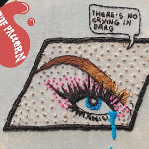Puede incluir: Ojo bordado con ceja marrón, iris azul y pestañas rosas y negras. El ojo está dentro de una forma delineada en negro, con el texto "THERE'S NO CRYING IN DRAG" en un bocadillo. El diseño está sobre tela clara.