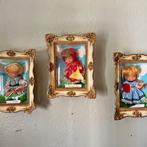 May include: Three framed pictures of dolls dressed in different costumes. The dolls are in a whimsical setting with a blue sky and clouds. The frames are gold with a white background. The text on the frames reads "Little Miss Muffet", "Red Riding Hood", and "Little Miss Muffet".