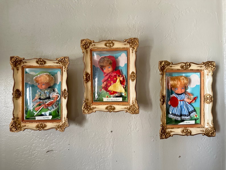 May include: Three framed pictures of dolls dressed in different costumes. The dolls are in a whimsical setting with a blue sky and clouds. The frames are gold with a white background. The text on the frames reads "Little Miss Muffet", "Red Riding Hood", and "Little Miss Muffet".