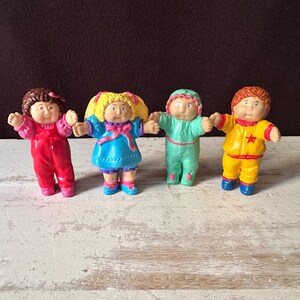 Op de afbeelding: Vier kleine plastic figuurtjes van kinderen met verschillende haarkleuren en kleding. Eén draagt een rode jumpsuit, één een blauwe jurk met geel haar, één een mintgroene onesie en één een gele outfit met een rode ster op de borst.